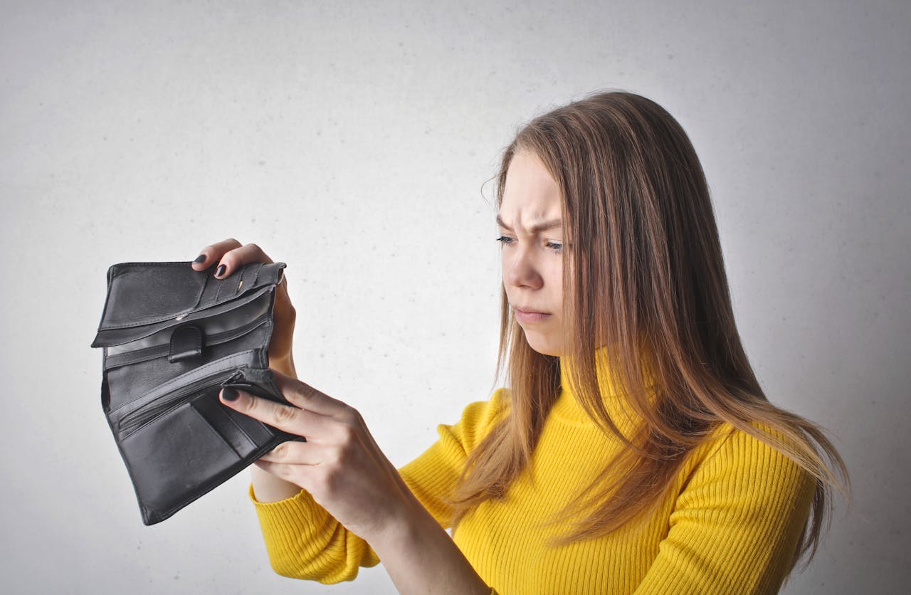 who-we-are Young woman in a yellow top looking at her empty wallet with a frustrated expression.